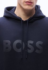 Εγγύς λήψη ατόμου που φοράει μπλε σκούρα κουκούλα με κορδόνια και τη λέξ η "BOSS" ανάγλυφα στο στήθος.