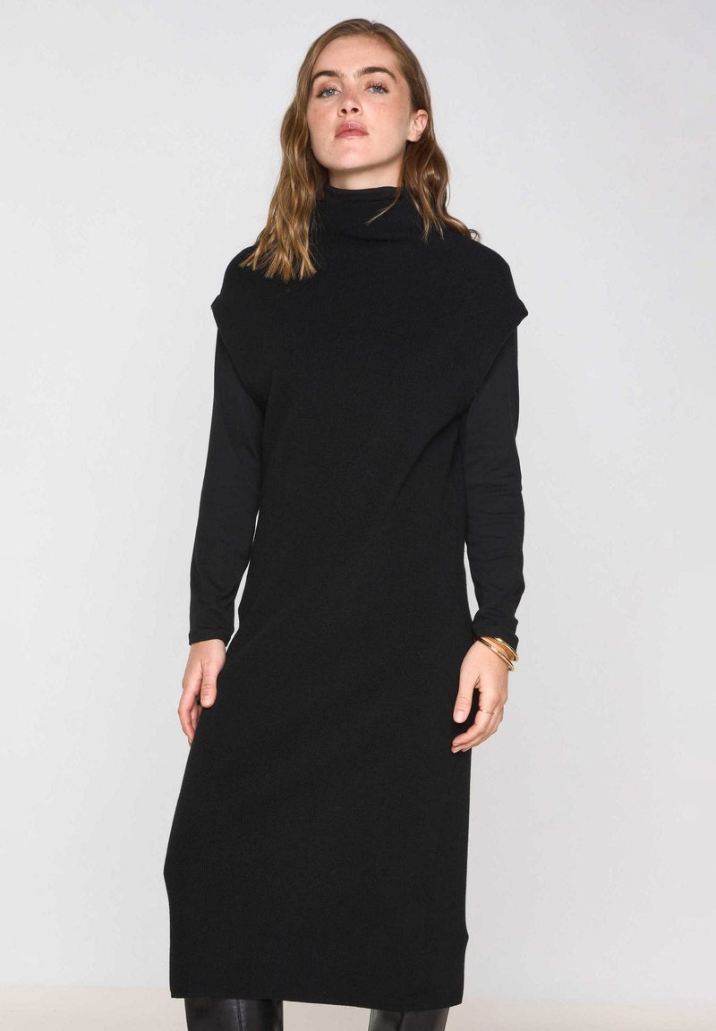 Abito lavorato a maglia nero con colletto alto, maniche lunghe e vestibilità ampia. Presenta un tessuto testurizzato e un design fino al ginocchio, senza motivi prominenti.