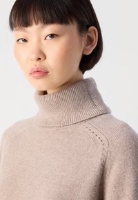 Giovane con capelli corti e scuri che indossa un maglione a collo alto in maglia beige, che guarda leggermente di lato su uno sfondo neutro.