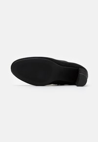 Zapato de mujer de ante negro tumbado de lado, mostrando la suela de goma texturizada y el tacón bajo en un fondo blanco.
