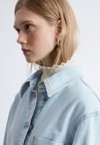 Light blue denim jacket with a folded collar, featuring contrast stitching and a button closure, layered over a cream frilled turtleneck.