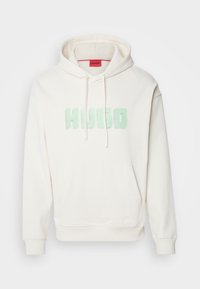 Sweat à capuche de couleur crème en tissu doux, avec une poche avant et un grand logo "HUGO" vert menthe sur la poitrine. Comprend une capuche ajustable par cordon.