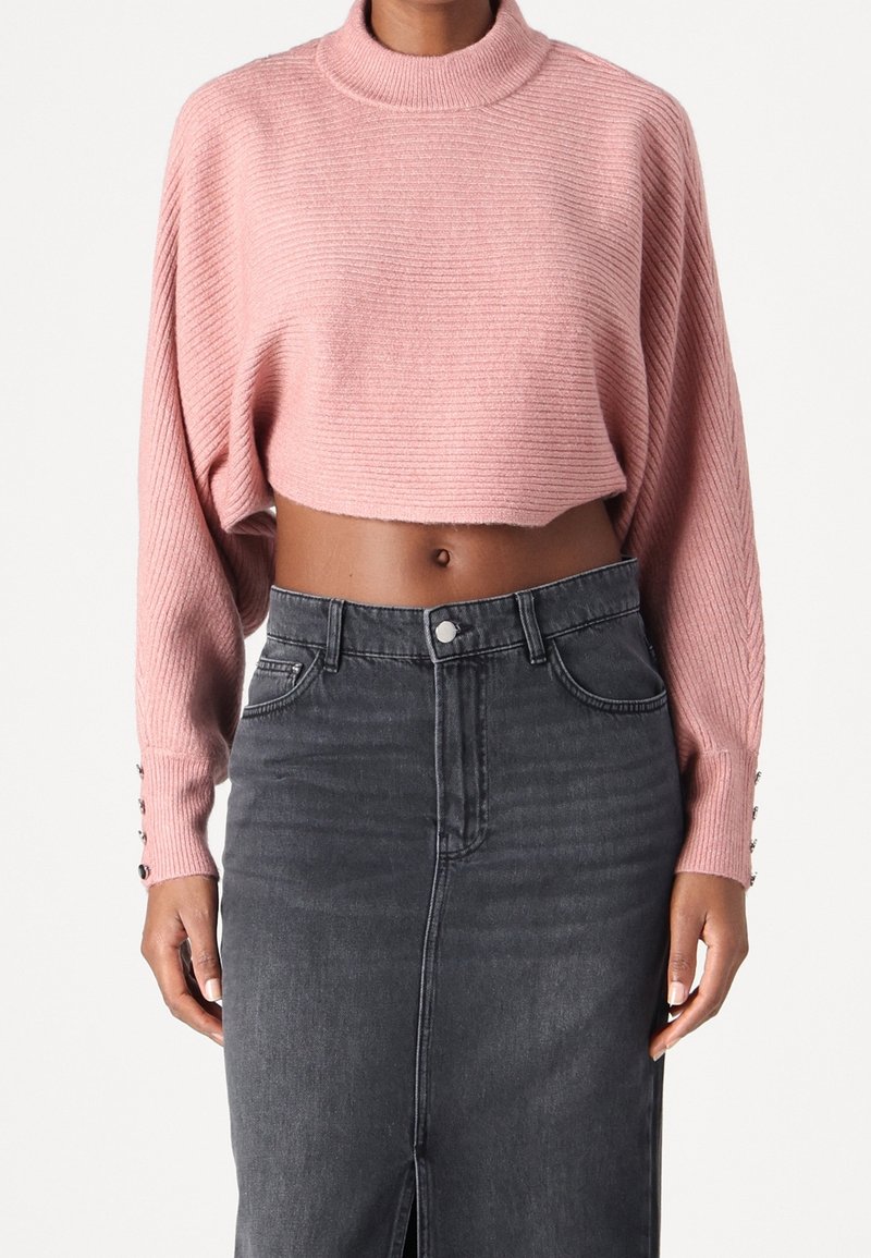 Femme portant un pull court rose côtelé avec des détails de boutons sur les manches et une jupe en jean gris foncé taille haute avec une fente à l'avant.