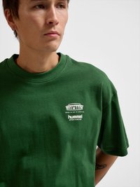 Młody mężczyzna z kolczykiem, ubrany w zieloną koszulkę Hummel Sportswear z białym logo i napisem na piersi, stojący na jednolitym tle.