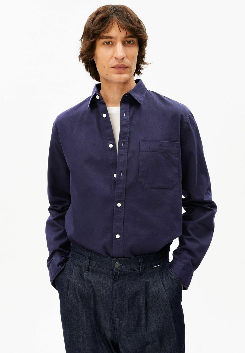 Homme aux cheveux mi-longs portant une chemise bleu foncé à boutons par-dessus un t-shirt blanc, les mains dans les poches d'un pantalon plissé foncé.