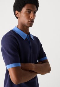 Mörkblå polo t-shirt med en texturerad väv, med en ljusblå krage och manschetter. Halvlånga ärmar, dragkedja fram och kontrasterande detaljer.