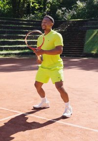 Tennis spiller iført en lys gul kortermet skjorte og shorts, holder en rød racket, på en leirkune med grønne omgivelser.