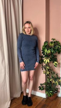 Jauna blondinė moteris vilki prigludusią mėlyną ilgomis rankovėmis suknelę ir juodus batus, stovinti šalia kambarinio marginto augalo vazonėlyje.