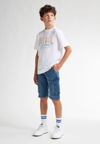 Camiseta blanca con "PETROL INDUSTRIES" impreso en dorado y verde. Pantalones cortos de carga azules con múltiples bolsillos. Zapatillas blancas y calcetines a rayas azules.
