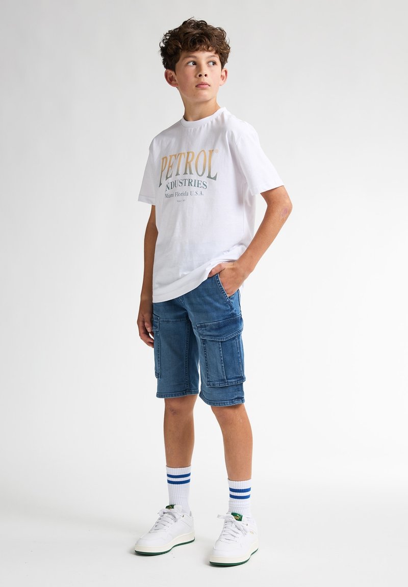 Camiseta blanca con "PETROL INDUSTRIES" impreso en dorado y verde. Pantalones cortos de carga azules con múltiples bolsillos. Zapatillas blancas y calcetines a rayas azules.