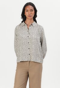 Beige bluse med prikkemønster i svart, med krage, knapplukking og lange ermer. Myk stoff med løs passform.
