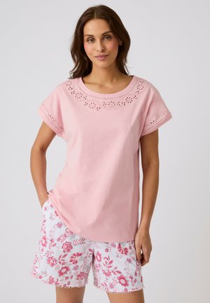 Vrouw draagt een roze top met korte mouwen met bloemuitstansingen en witte korte broek met roze bloemmotieven, staand tegen een effen achtergrond.