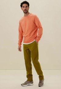 Koraal sweatshirt, ribbelmanchetten, witte binnenlaag; olijfgroene broek, rechte snit; grijze sneakers met witte accenten. Neutrale achtergrond.