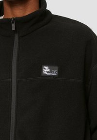 Schwarze Fleecejacke mit hohem Kragen, Reißverschluss und kleinem Markenpatch auf der Brust. Weiche Textur und minimalistisches Design.