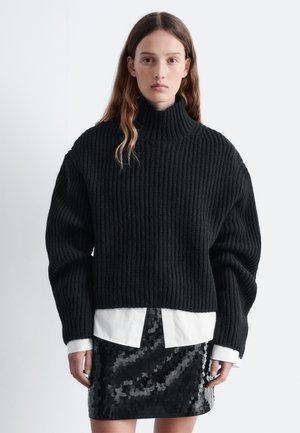 Schwarzer gerippter Pullover mit hohem Kragen und übergroßen Ärmeln, kombiniert mit einem weißen Button-Down-Hemd und einem schwarzen Glitzerrock.