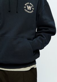 Navy blue sweatshirt with a front pocket, featuring a circular logo in light beige text. It has a drawstring hood and ribbed cuffs.