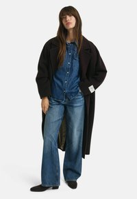Un long manteau sombre sur une chemise en denim bleu et un jean large bleu. Bottines noires. Textures lisses, couleurs contrastées et design structuré.