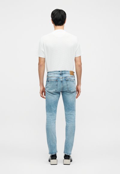 Ανοιχτό μπλε τζιν από τζιν ύφασμα, εφαρμοστή σχεδίαση, στυλ με πέντε τσέπες, με καφέ δερμάτινο ταμπελάκι στο πίσω μέρος; φορεμένα με απλό λευκό μπλουζάκι.