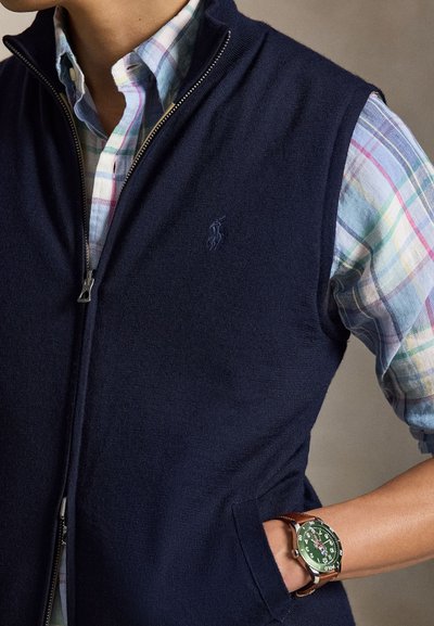 Vīrietis valkā tumši zilu rāvējslēdzēja vestīti virs daudzkrāsaina pundura krekla ar zaļu ciparnīcas pulksteni un brūnu ādas siksniņu.