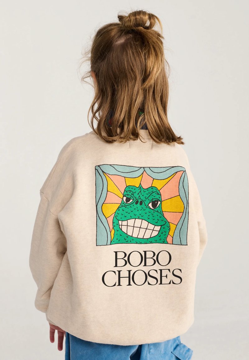 Enfant aux cheveux châtain clair attachés en demi-chignon porte un sweat-shirt beige avec une grenouille colorée souriante et le texte "BOBO CHOSES" dans le dos.