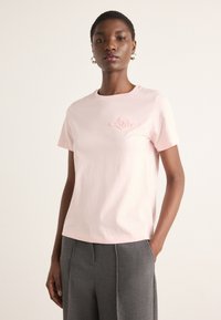 Helles rosa Baumwoll-T-Shirt mit Rundhalsausschnitt, kurzen Ärmeln und einem gestickten Logo in ähnlichen Farbtönen auf der Brust. Getragen mit grauen Hosen.