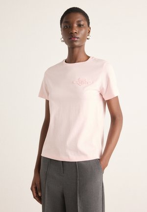 T-shirt in cotone rosa chiaro con collo a giro, maniche corte e un logo ricamato in tonalità tonali sul petto. Indossato con pantaloni grigi.