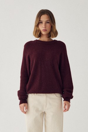 Žena s vlasmi do ramien, oblečená v voľnom vínovom svetri a svetlo béžových nohaviciach, stojaca pred jednofarebným pozadím.