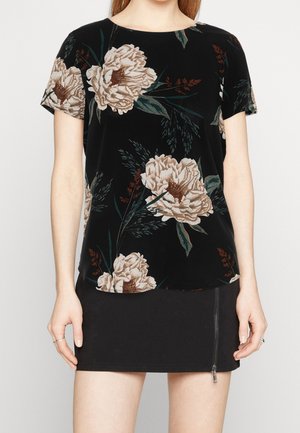 Vrouw draagt een zwart kortemouwtop met grote beige bloemenprint en een zwarte rok met een ritssluiting aan de voorkant.