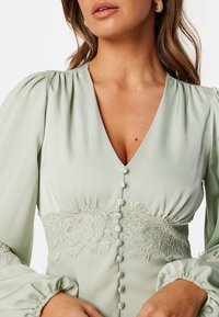 Světle zelená blůza s hlubokým V-výstřihem, nabíranými rukávy, krajkovými detaily v pase a knoflíkovými akcenty na přední strani.