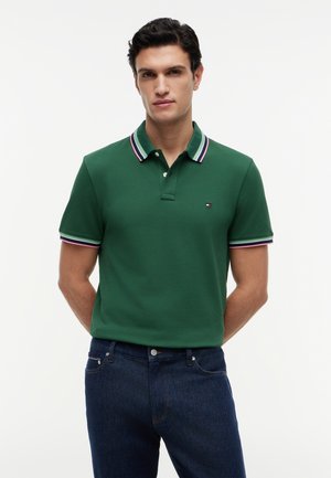 Mand med mørkt hår står og vender fremad iført grøn polo-shirt med stribet krave og mørkeblå jeans mod en ensfarvet baggrund.