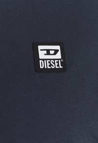 Mörkblått tyg med en svart fyrkantig lapp som har en vit Diesel-logotyp och stiliserat "D"-emblem i mitten.