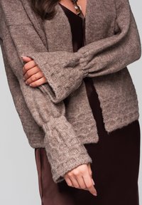 Brauner, oversized Strickcardigan mit strukturierten Ärmeln, offener Vorderseite und weichem Finish, getragen über einem burgunderfarbenen Kleid, das einen geschichteten Look zeigt.
