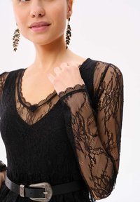 Zwarte kanten jurk met lange doorzichtige mouwen met bloemmotieven, een V-halslijn en een zwarte riem met een zilveren versierde gesp.