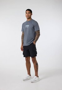 Tatuiruotes turintis vyras vilkintis pilką marškinėlį su piešiniais, juodas cargo šortas ir baltus sportbačius, stovintis prieš šviesų vientisą foną.