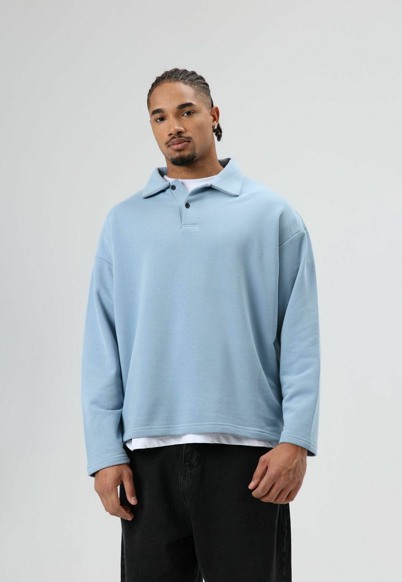 Hellblauer Pullover mit Kragen, zwei Knöpfen und langen Ärmeln. Weicher Stoff mit lockerem Sitz, getragen über einem weißen Hemd und schwarzen Hosen.