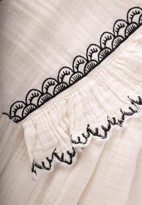 Tissu blanc avec un motif festonné brodé en noir et une texture froncée le long du bord, créant un détail décoratif.