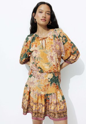 Robe florale multicolore avec des manches bouffantes, un col à nœud et un ourlet en couches. Le tissu présente des motifs complexes dans des teintes orange, verte et violette.