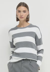 Donna con capelli corti castani che indossa orecchini a cerchio argentati, un maglione a righe grigie e bianche, e pantaloni grigi con le mani in tasca.