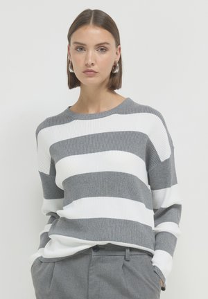 Femme aux cheveux bruns courts portant des boucles d'oreilles en argent, un pull à rayures gris et blanches, et un pantalon gris avec les mains dans les poches.