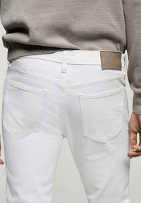 Białe dżinsy z denimu o teksturowanym wykończeniu, z dwiema tylnymi kieszeniami i brązową skórzaną metką na pasku. Prosty i schludny design.