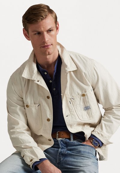 Ανοιχτό καφέ σακάκι με κουμπιά, διαθέτει μικρή τσέπη στο στήθος και διακριτά ραφές. Φοριέται πάνω από ένα ναυτικό μπλουζάκι, συνδυασμένο με μπλε τζιν.