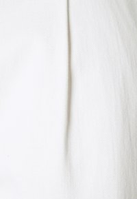 Vitt tyg som visar en texturerad, diagonalt väv. En vertikal söm löper längs ena sidan, vilket ger dimensionell detalj till textilen.