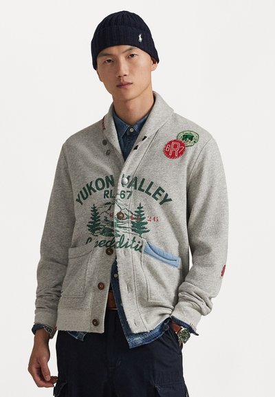Cardigan gris avec impression graphique verte, poche bleue et boutons marron. Porté par-dessus une chemise en jean, associé à un bonnet en tricot bleu marine.