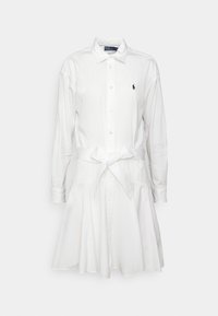 Biała bawełniana sukienka koszulowa z długimi rękawami, zapinana na guziki z wiązanym paskiem w talii. Ma małe czarne logo na górnej lewej stronie.