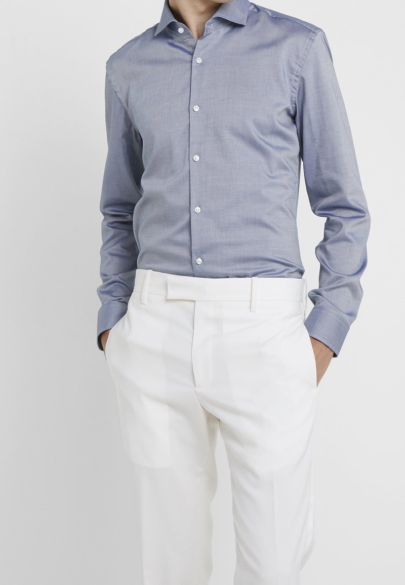 Homme portant une chemise bleu clair près du corps boutonnée, rentrée dans un pantalon blanc sur mesure, les mains dans les poches, sur un fond uni.
