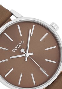 Montre à cadran marron avec des index et aiguilles argentés. Présente une lunette en argent et un bracelet en cuir marron. Design minimaliste.