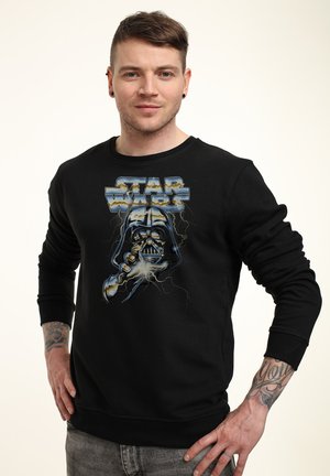 Hombre con cabello corto que lleva una sudadera negra de Star Wars con Darth Vader y relámpagos, de pie con las manos en las caderas.