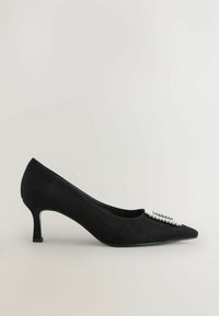 Escarpin en suède noir avec un bout pointu et un accent décoratif en rhinestones. Le talon est fin et mesure environ deux pouces de hauteur.