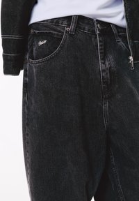 Lähellä otettu kuva henkilöstä, joka wears mustat denim-farkut ja musta takki, jossa on valkoiset saumat, esittäen etutaskuja ja vetoketjuyksityiskohtia.