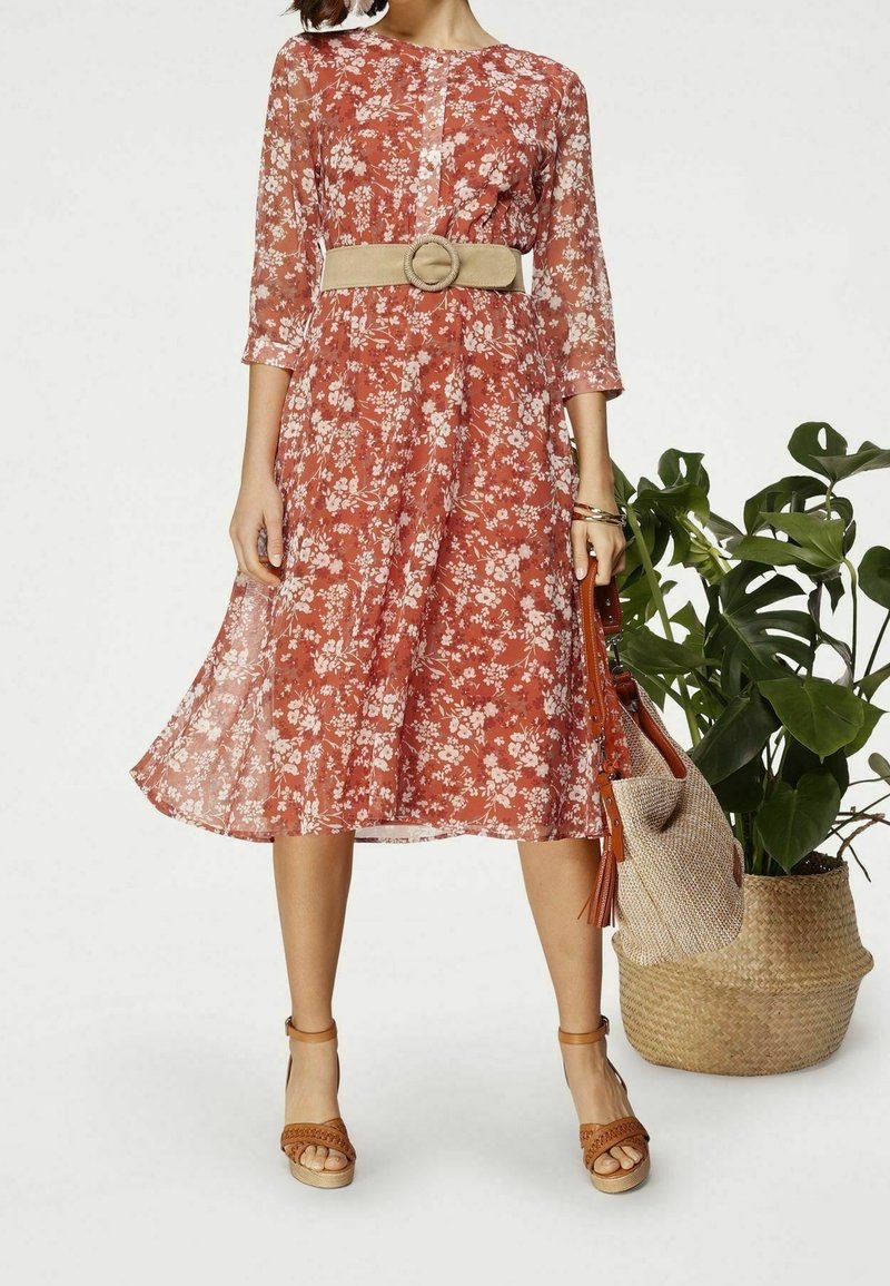 Femme portant une robe midi fleurie couleur rouille avec une ceinture beige et des sandales compensées marron, tenant un sac tissé à côté d'une grande plante verte en pot.
